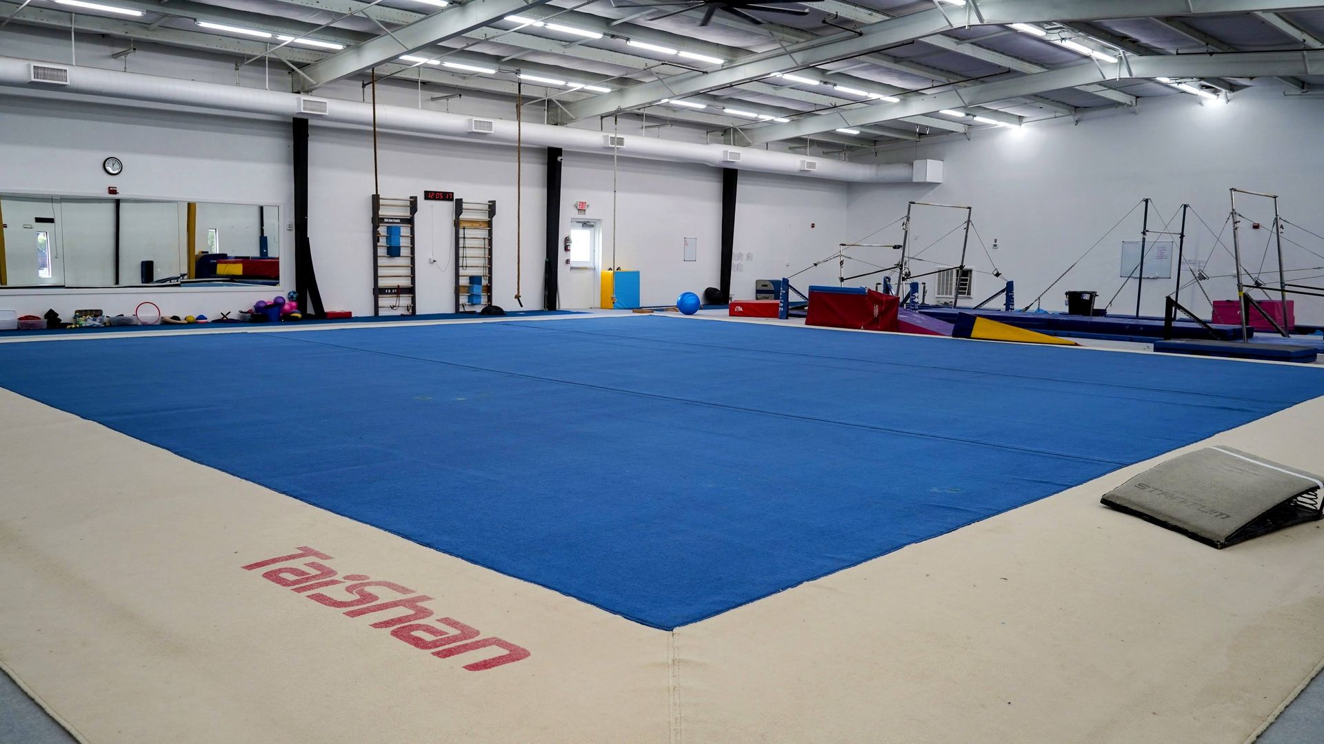 Large indoor gymnastics gym with blue floor mat, ramps, and equipment under bright overhead lights.
