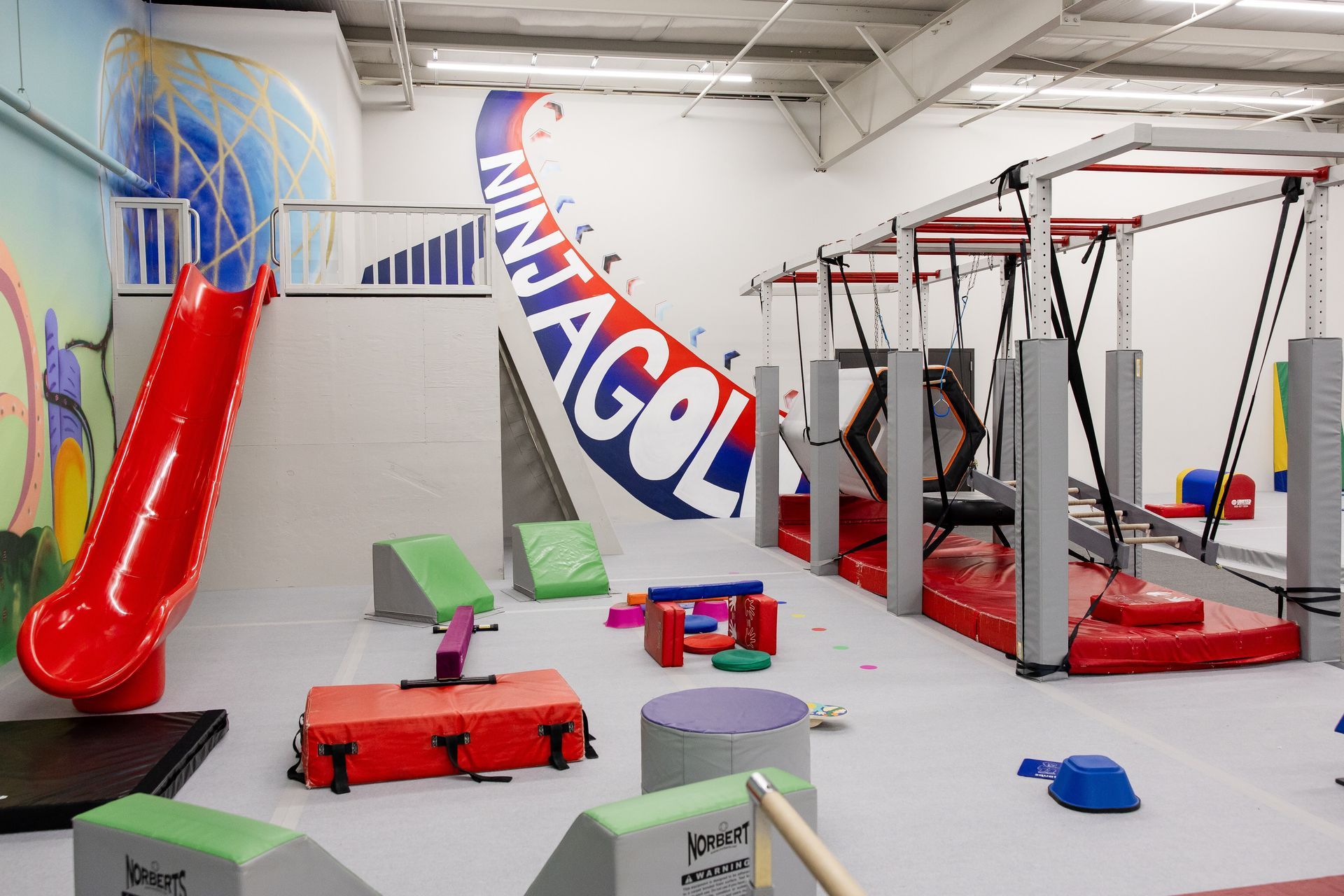Indoor playroom with red slide, climbing structure, mats, and colorful foam seating under a “Ninja Gold” mural