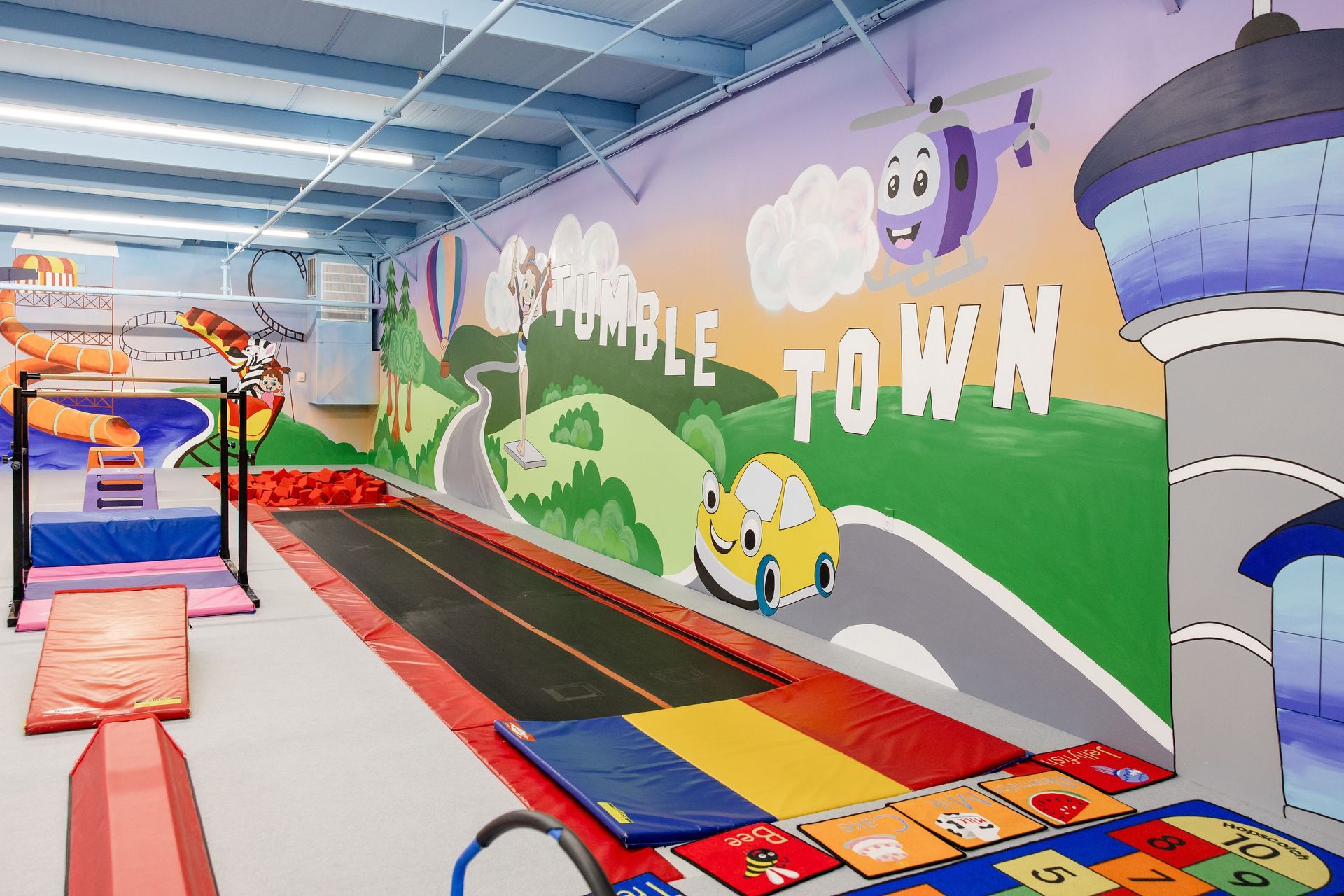 Colorful indoor play area with a trampoline, mats, and a cartoon mural reading “Tumble Town”