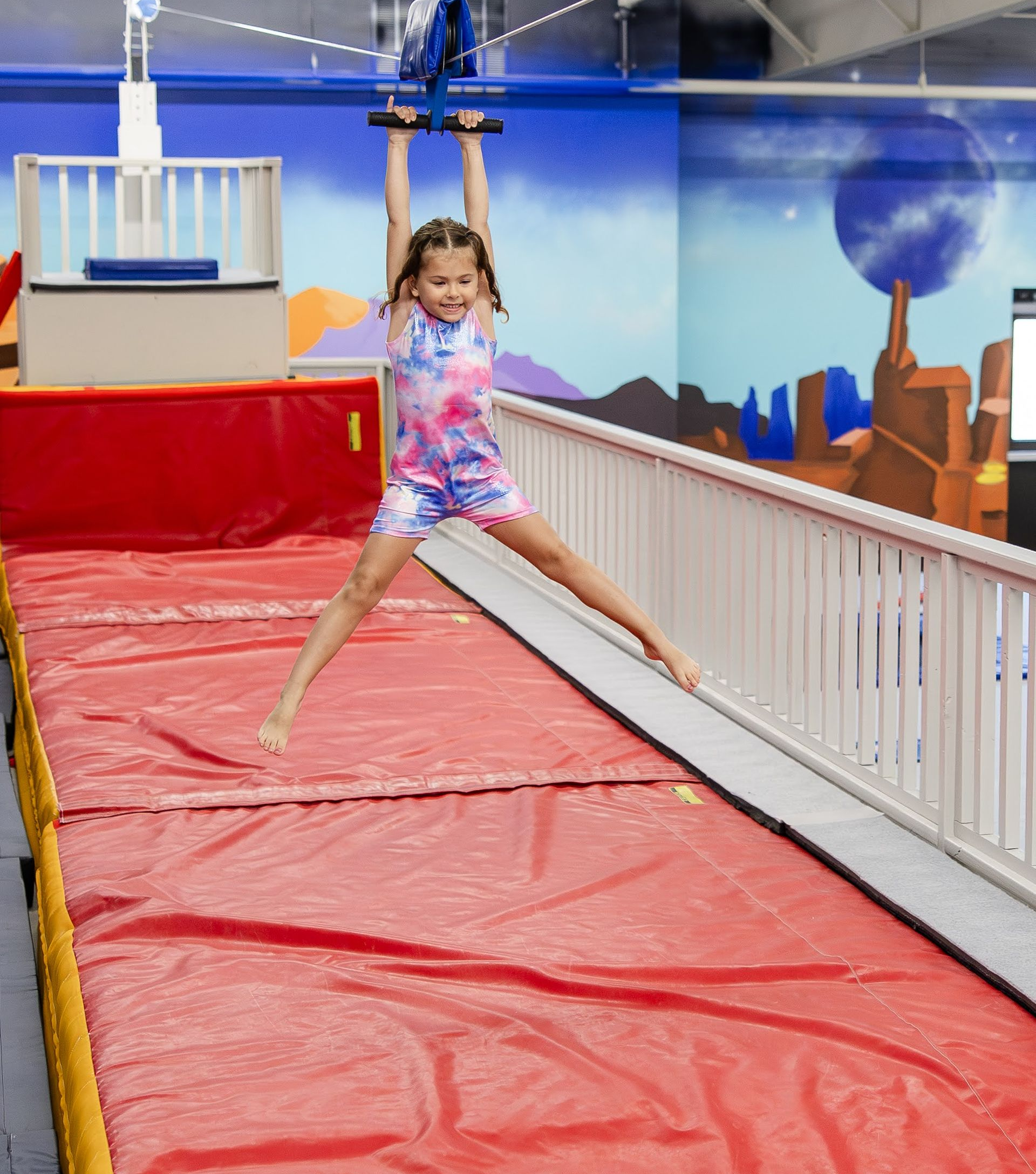 Child in colorful leotard hanging from zipline above a red padded gym mat, legs spread wide.