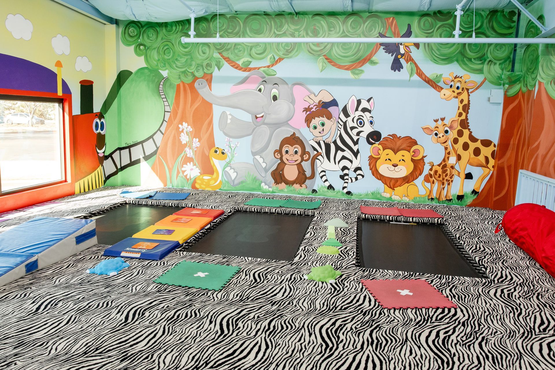 Colorful indoor kids playroom with zebra rug, animal mural, trampolines, and soft play equipment