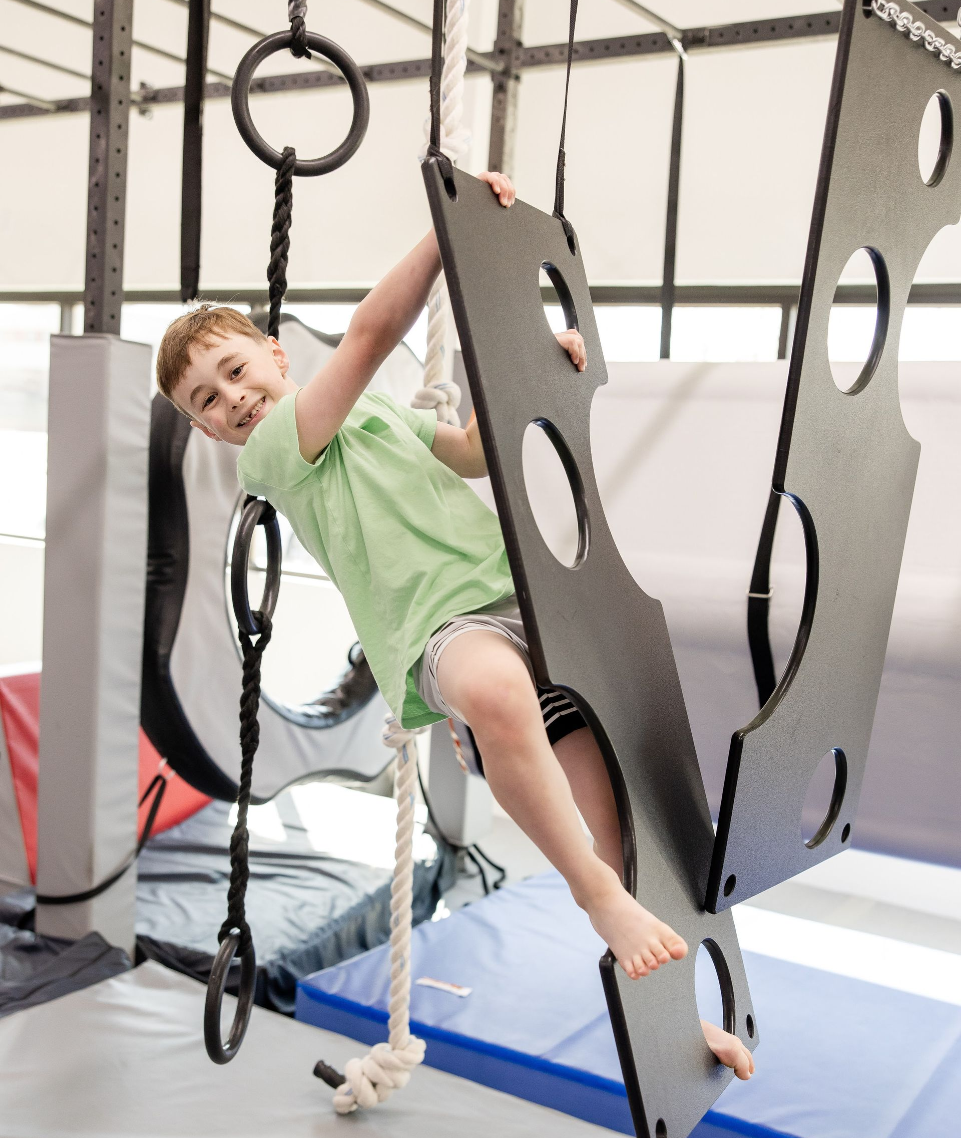Child climbing on hanging ninja playground panels in an indoor gym, smiling and wearing a green shirt