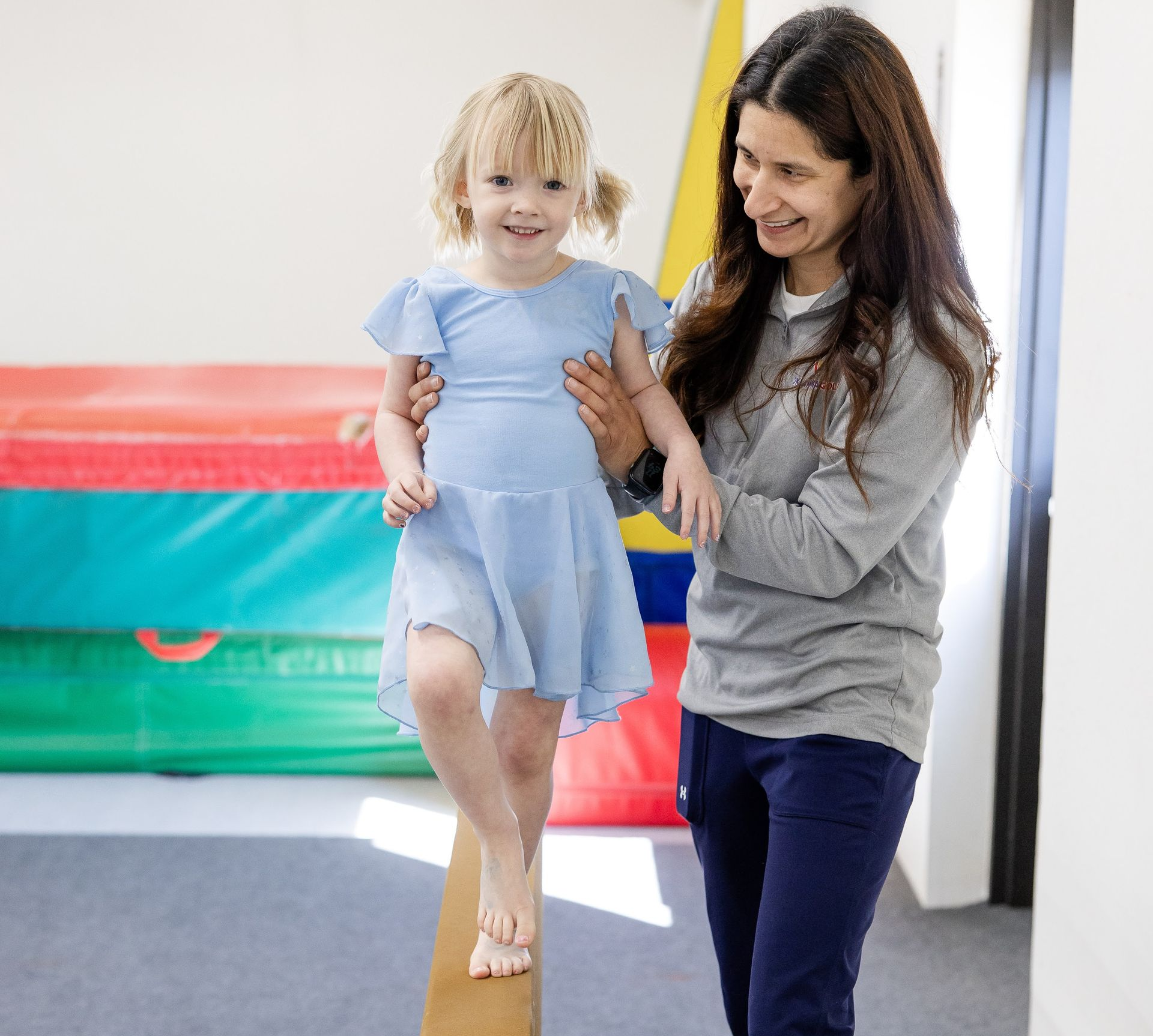 Woman helping child balance on a beam in a bright playroom