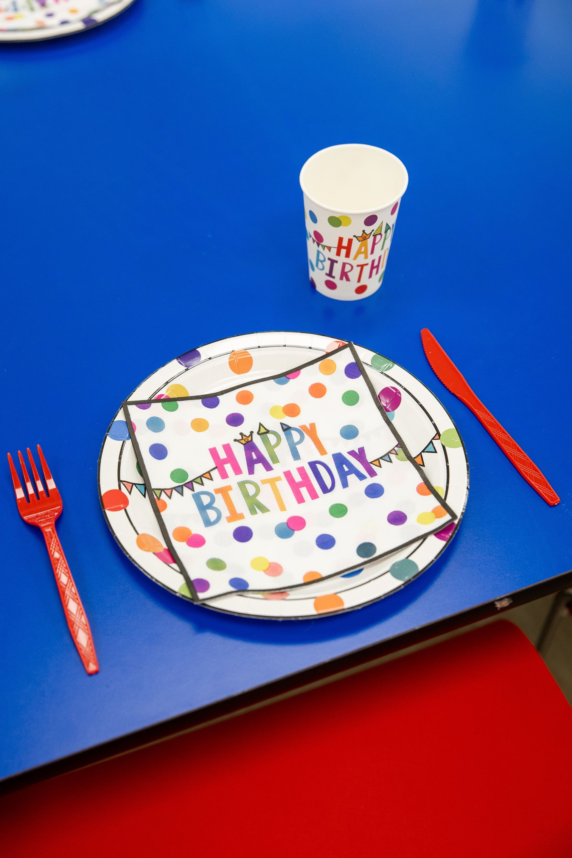 Birthday table setting with colorful “Happy Birthday” plate, cup, fork, and red knife on blue tablecloth