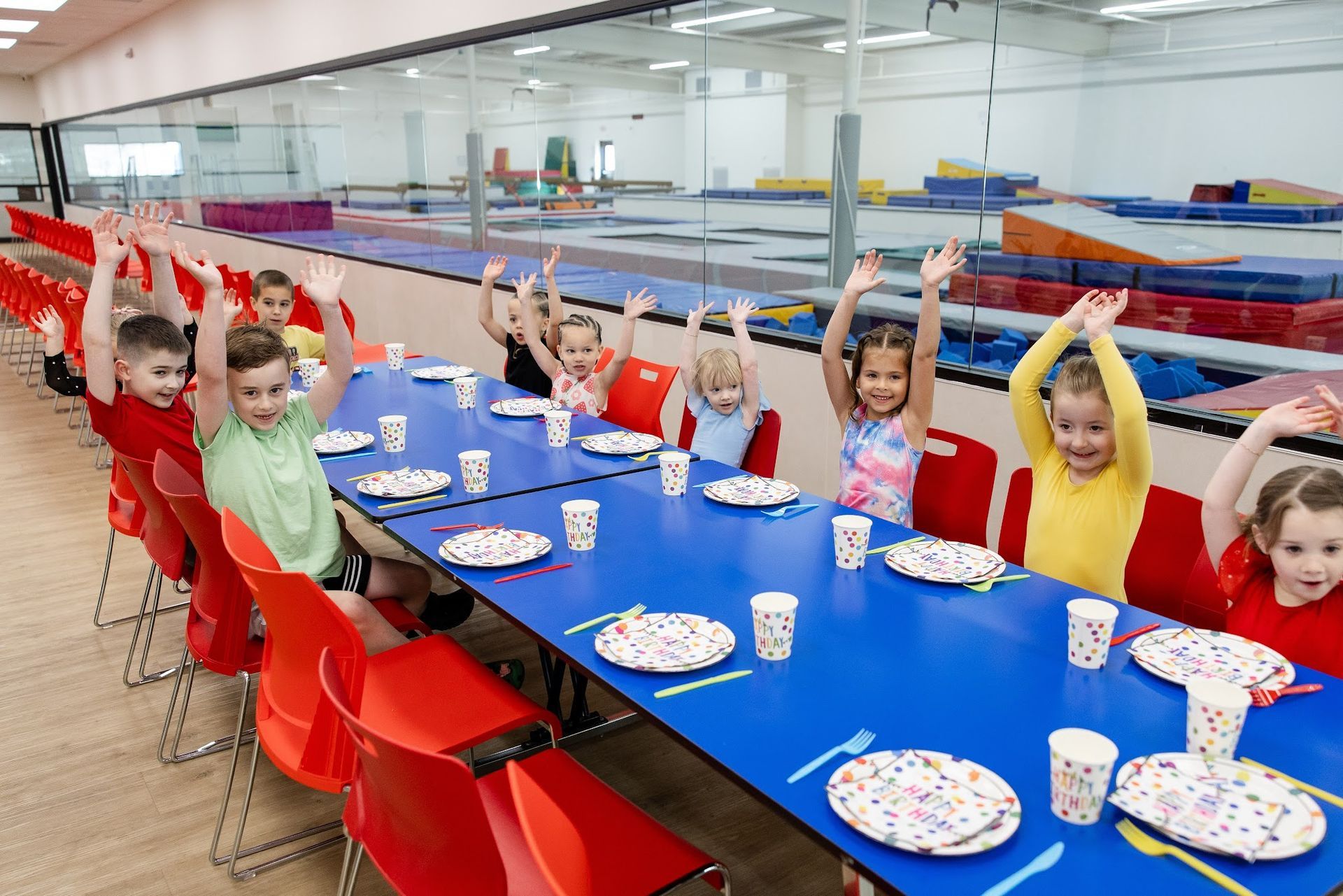 Children seated at a long blue table in a bright cafeteria, raising their arms during a birthday party.
