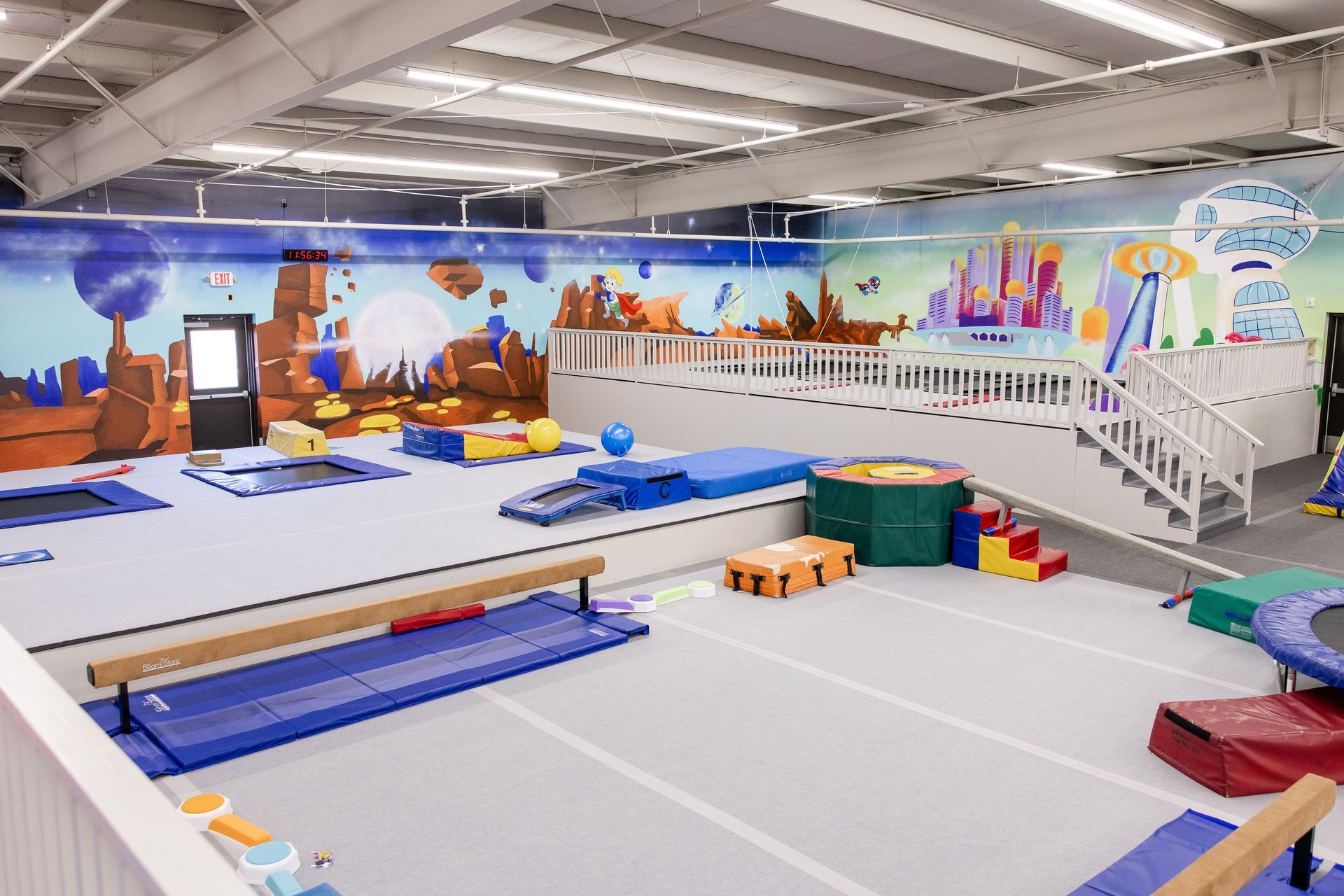 Indoor gym with mats, balance beams, foam blocks, and a colorful wall mural
