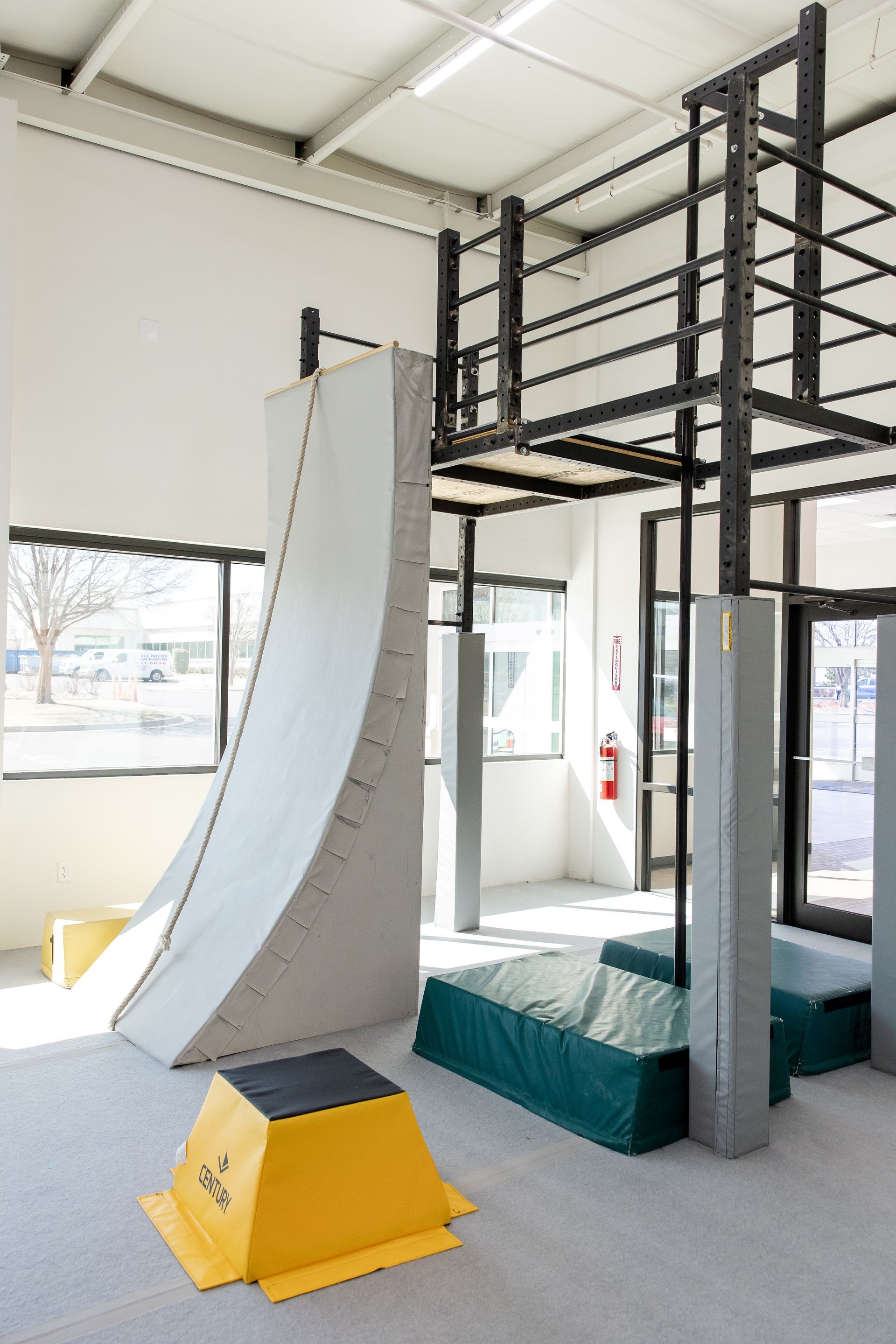 Indoor climbing wall with a curved gray ramp, black platform above, and padded landing mats