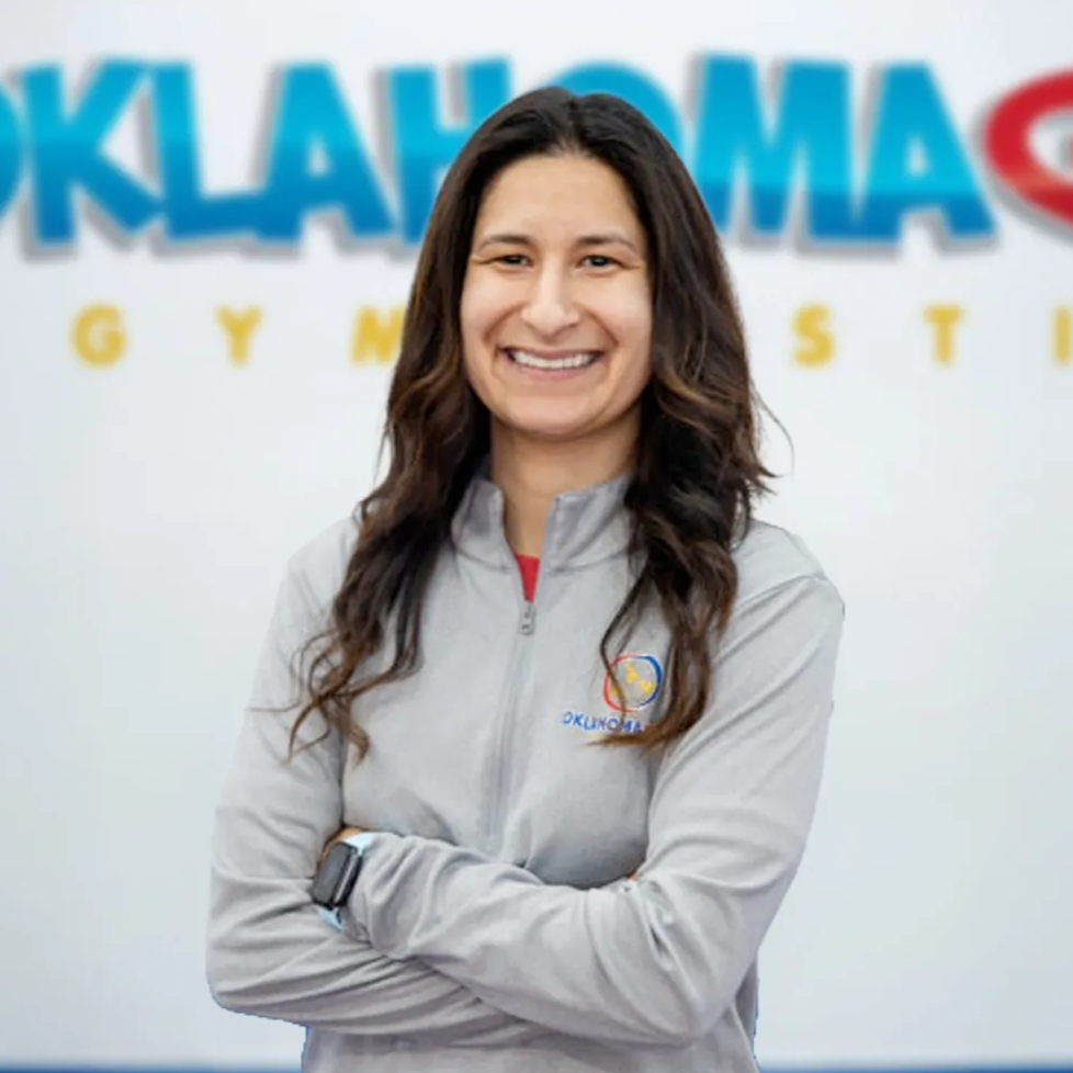 Smiling staff member in a gray jacket standing before an Oklahoma Gymnastics sign