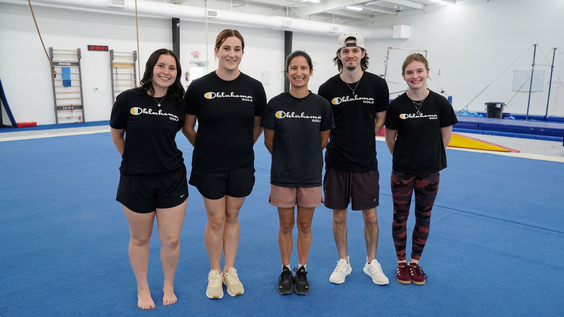 Five gymnastics coaches in black shirts standing on a blue gym mat inside an indoor training facility.