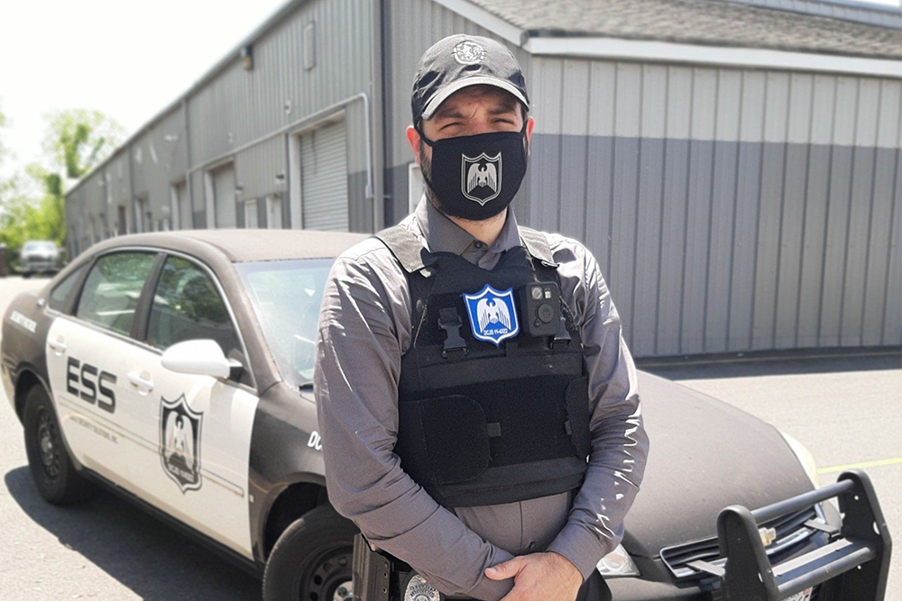 Man in uniform wearing a mask and body armor stands in front of a security car.