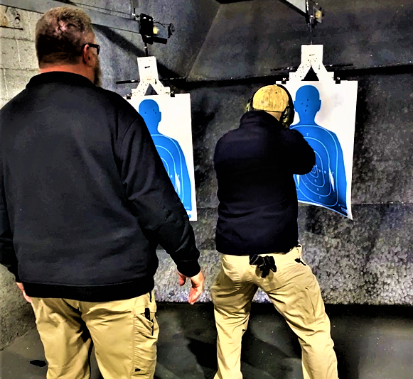 Two men at an indoor shooting range; one aims a pistol at a blue silhouette target, the other observes.
