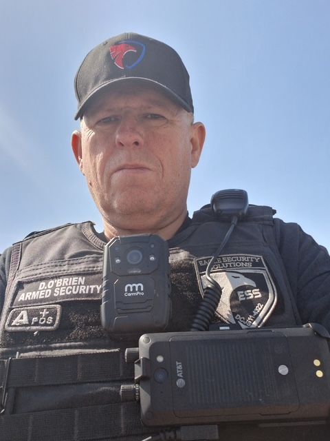 A security officer in a black uniform and cap with a body camera and radio, looking directly at the camera.