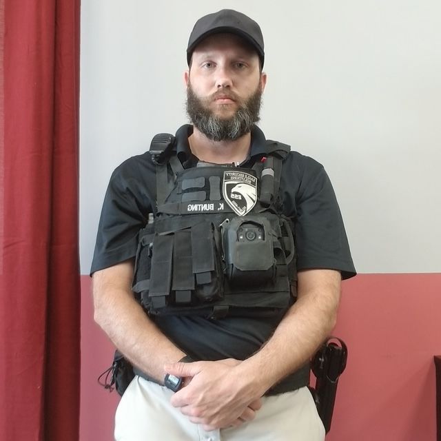 A person with a beard, wearing a black cap, polo shirt, and tactical vest, stands with hands clasped against a wall.