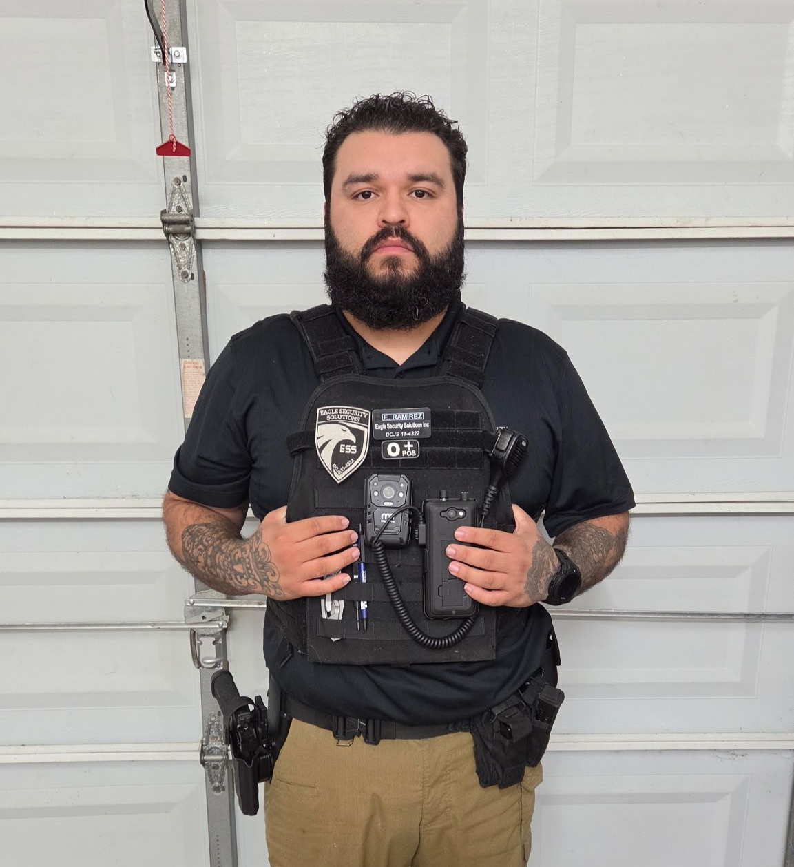 A bearded individual in a black polo shirt and tan pants wears a tactical vest with gear against a garage door background.
