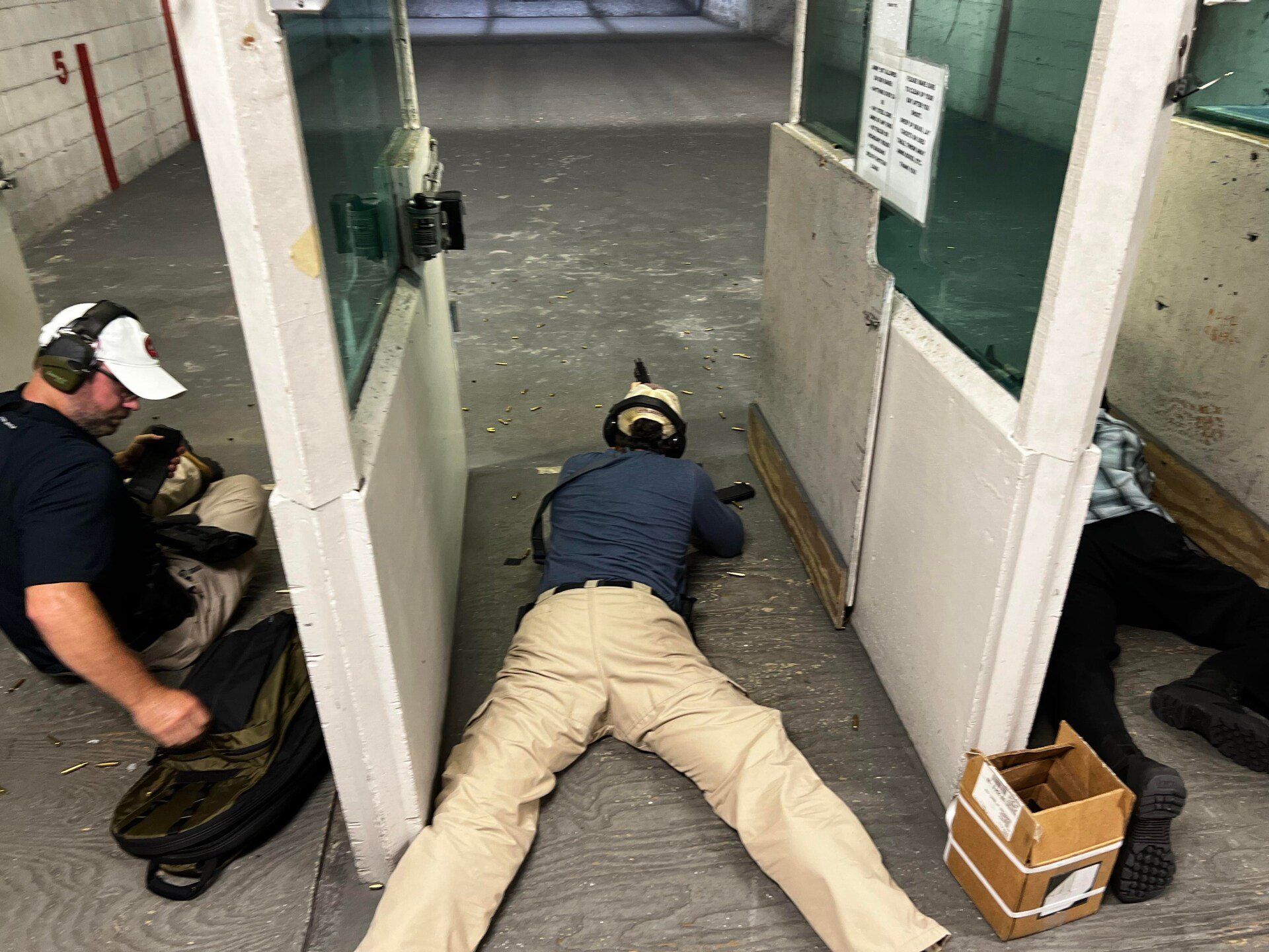 Person aiming a rifle at a shooting range, lying down with legs spread. Two other people are present.