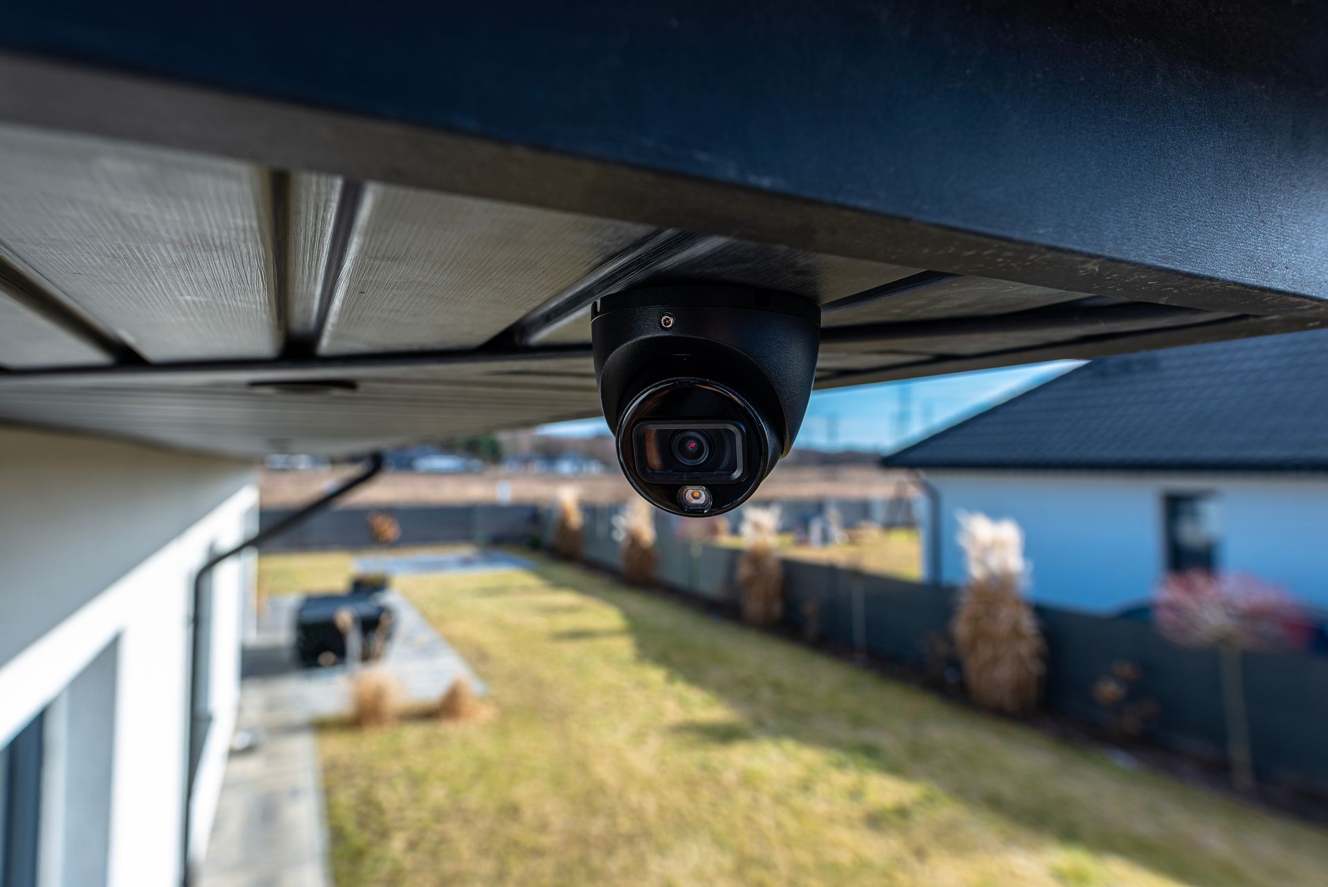 Black dome residential security systems camera mounted under a roof eave overlooking a backyard.