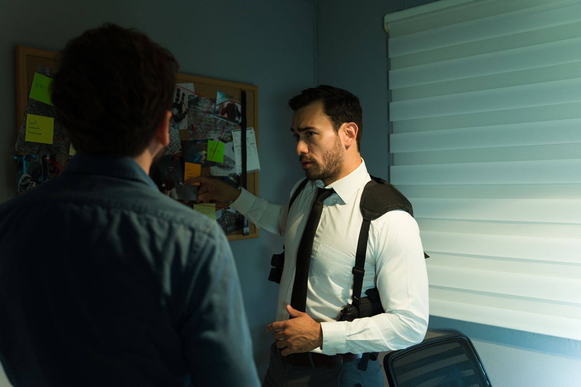 Private investigator pointing to an evidence board while briefing a client in a dim office Private investigator pointing to an evidence board while briefing a client in a dim office