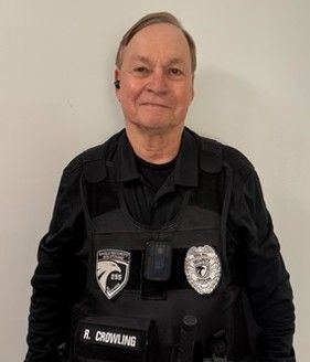 A security guard in a black uniform and tactical vest with a badge and name tag stands against a plain white wall.
