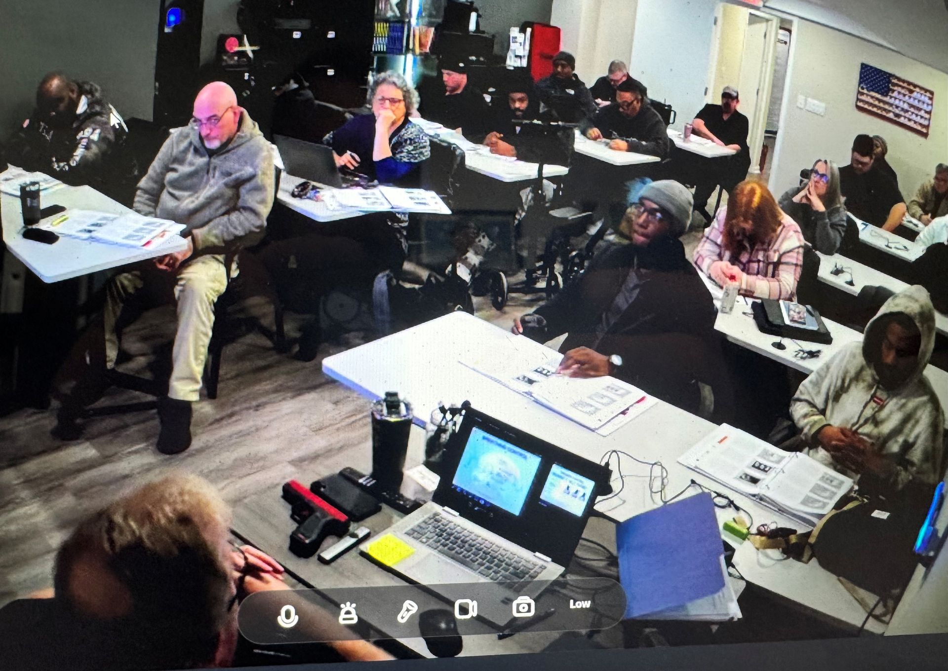 Classroom setting with diverse people seated at desks, some looking at materials, an American flag in the background.