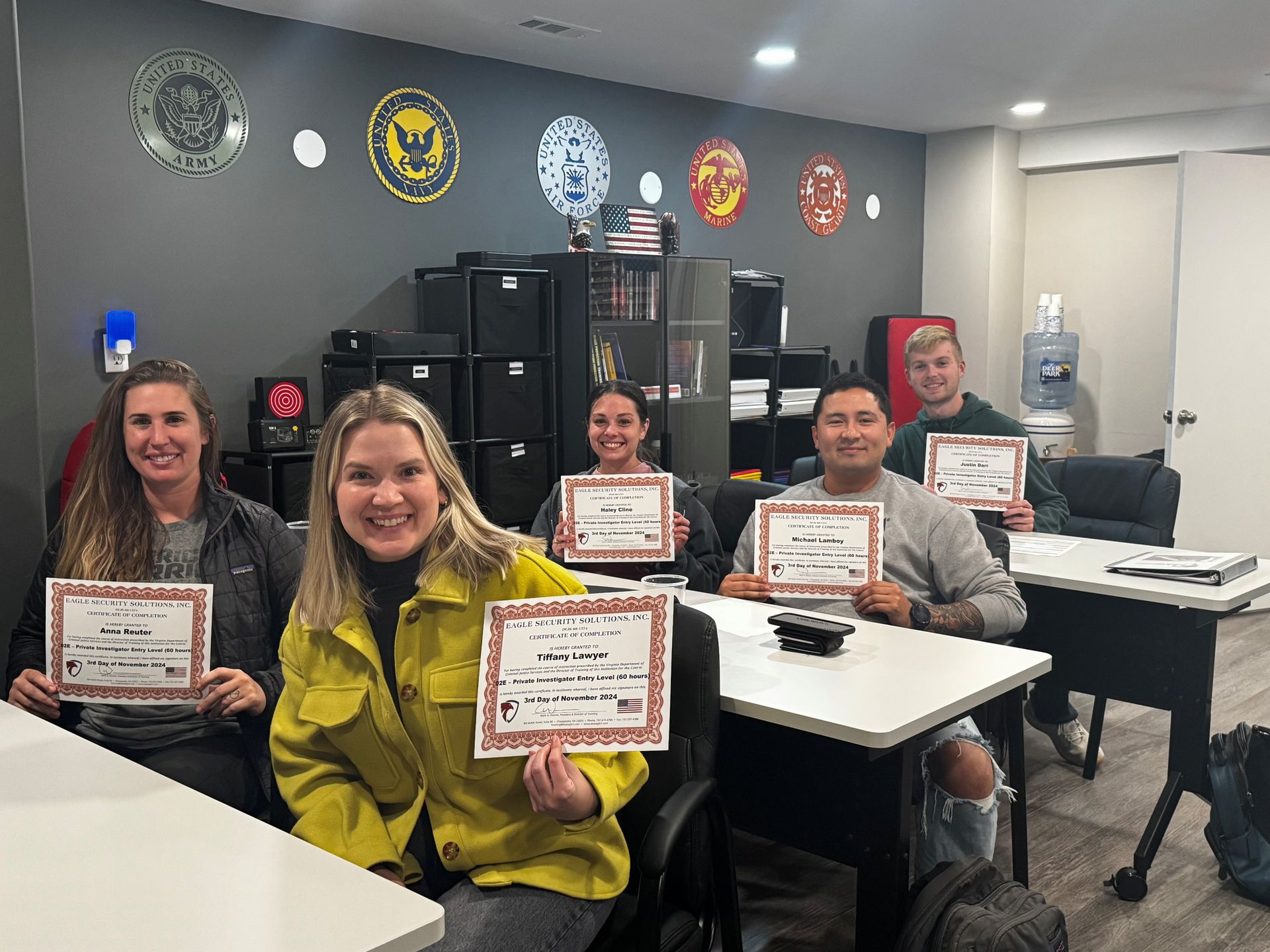 People smiling, holding certificates, in a classroom. Walls have military-themed logos.