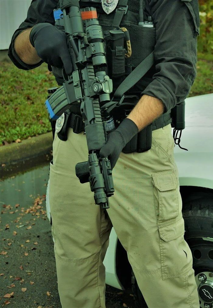 Police officer in tactical gear holding a rifle, outdoors near a vehicle.