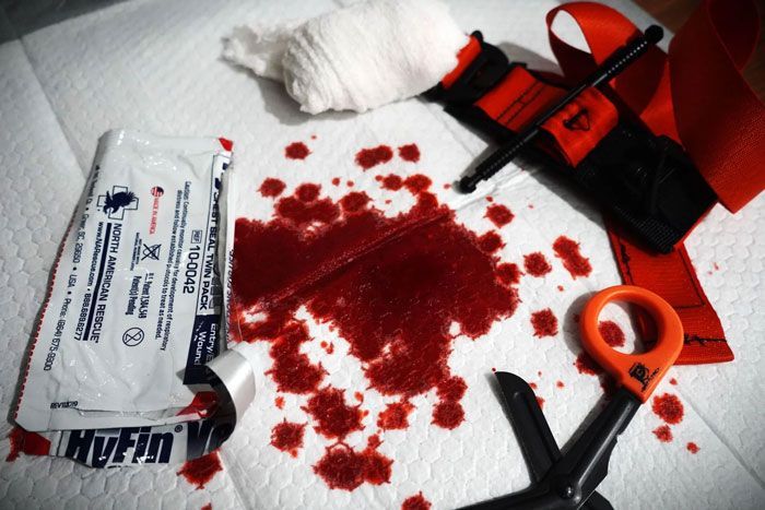Blood, gauze, and a tourniquet on a white surface, with medical shears and a bandage packet.