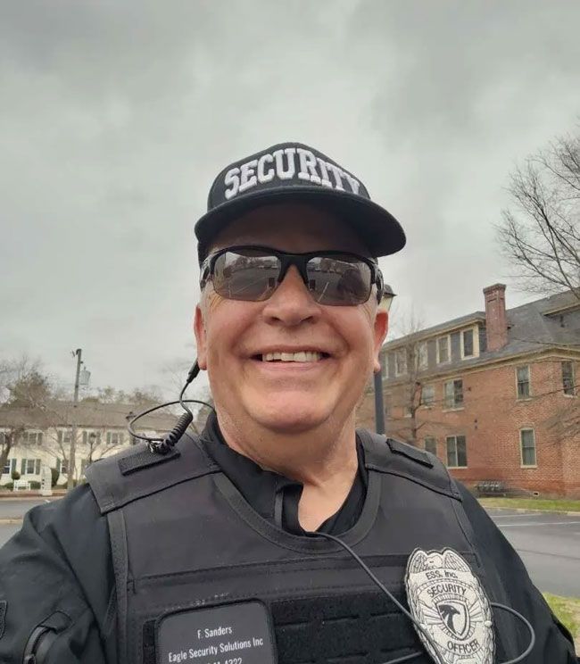 Security guard in uniform wearing a cap and sunglasses, smiling in front of a building.