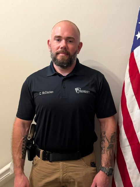 A man in a black polo shirt and khaki pants stands in front of an American flag, wearing a holstered firearm.