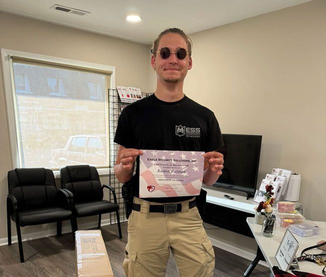 Man in black shirt and tan pants holds a certificate, standing in an office.