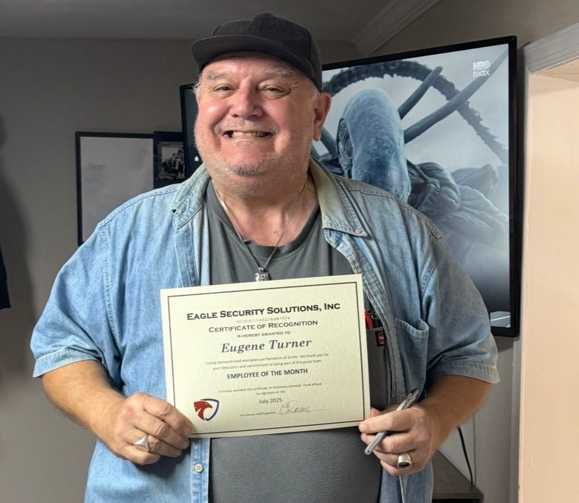 Man holding certificate, smiling. Wearing denim shirt, cap, necklace. Background: artwork, framed photo.