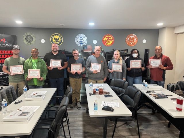Group of people holding certificates, inside a room. Tables in foreground, military emblems on the wall.