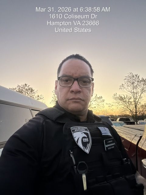A security officer in a black uniform and vest standing outdoors at sunrise, with a vehicle in the background.