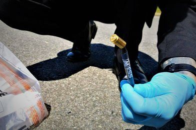 Person wearing a blue glove holding a vial. The person appears to be crouching on pavement.