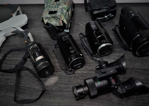 Various cameras and camcorders on a dark wooden surface. Includes a drone and a night vision device.