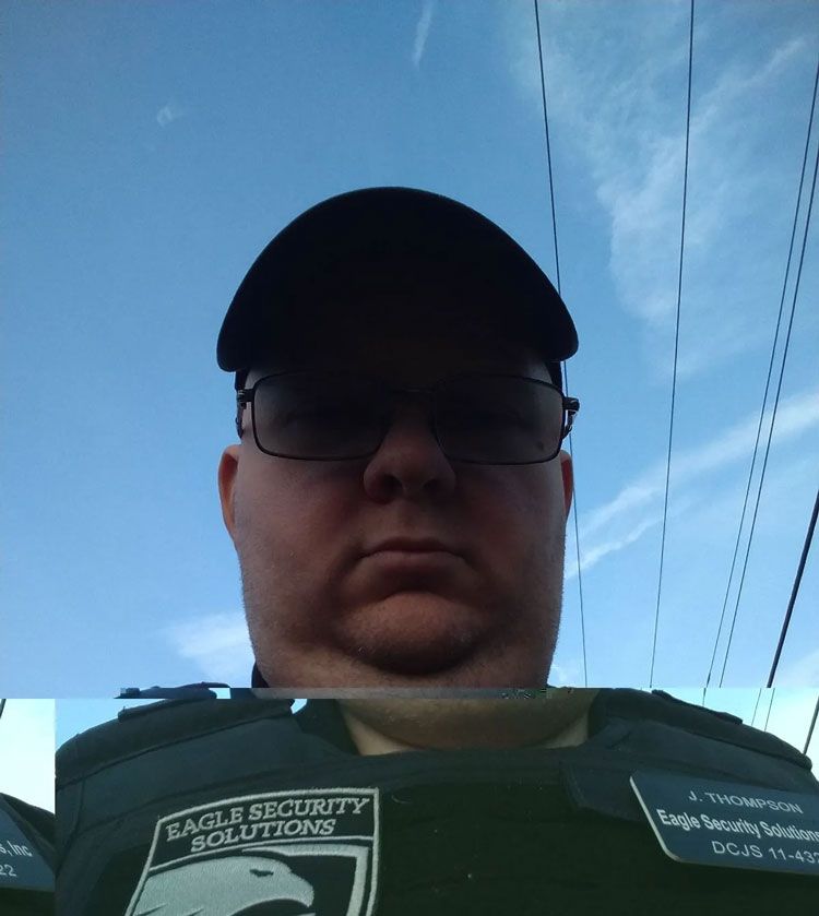 Man wearing glasses and a hat, in a security uniform, against a blue sky. The uniform has