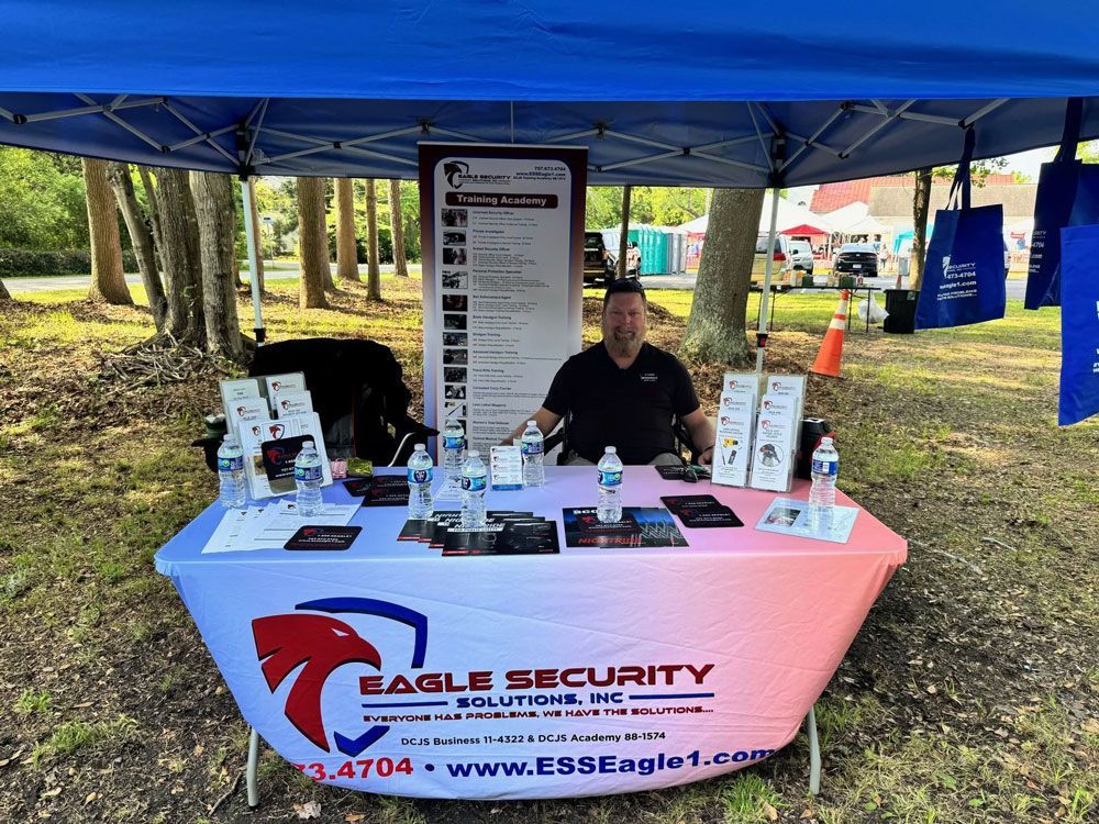 Man at Eagle Security Solutions booth under a tent. Table displays products, sign, and water bottles. Outdoor setting.