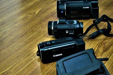 Three black camcorders on a wooden surface, one Canon and two Sony models, with a partially visible black charging device.