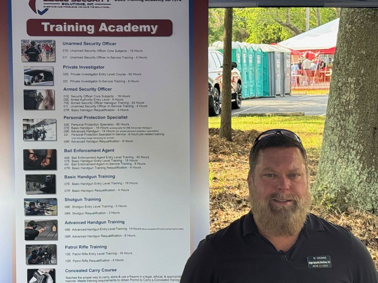 Man stands by a training academy banner outdoors, smiling with a beard; portable toilets visible in background.
