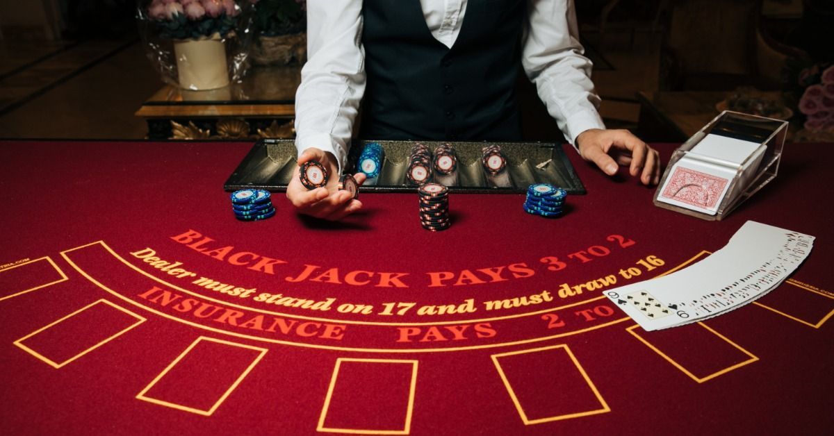 A dealer holds casino chips over a red blackjack table as playing cards and stacked chips cover the 