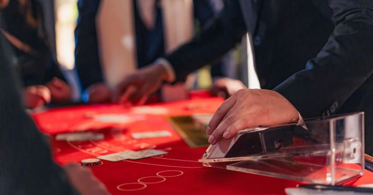 A dealer handles a card shoe at a red casino table while guests in formal attire gather around.