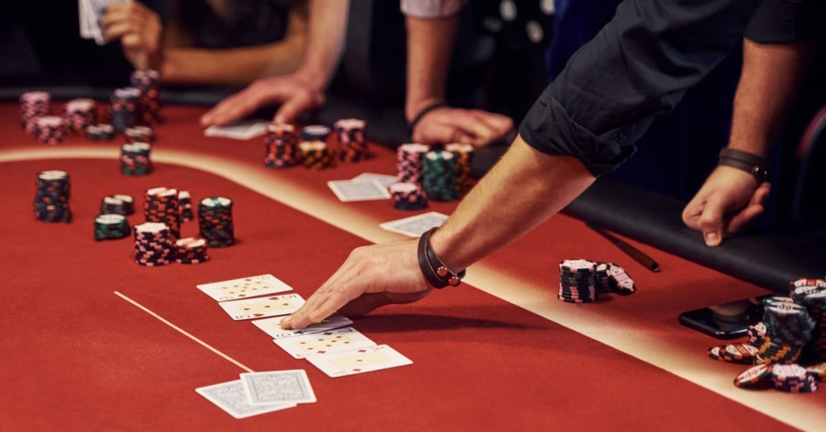 A man wearing a black shirt touches the board on a red poker table as guests sit behind stacks of colorful casino chips.