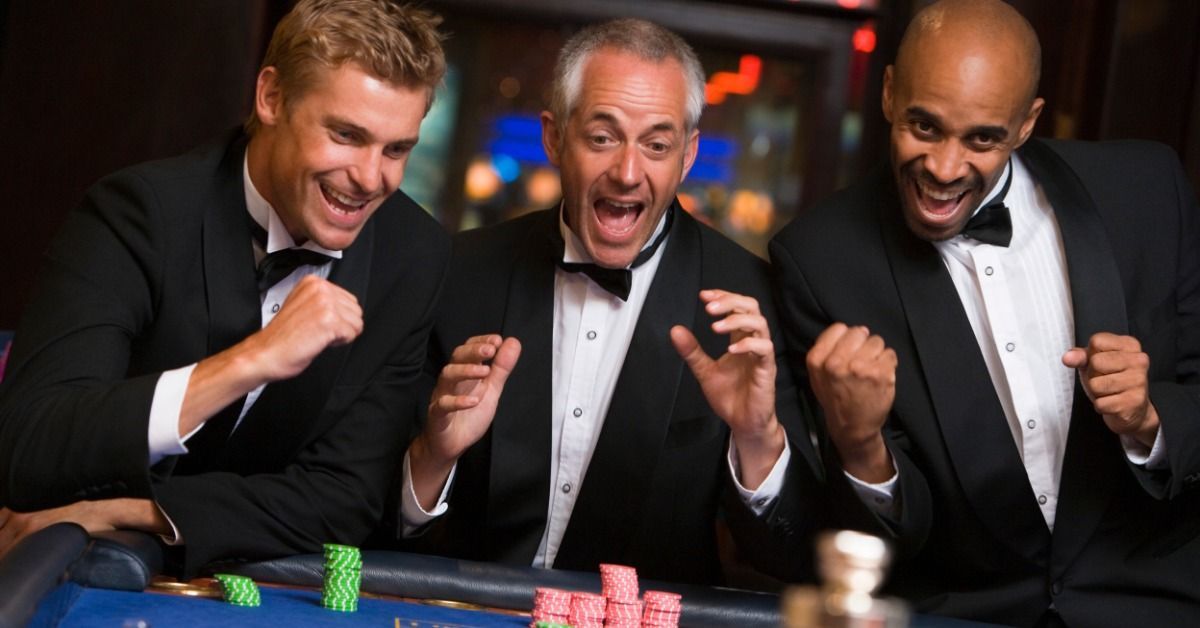 Three men in tuxedos cheer around a blue roulette table while colorful casino chips cover parts of the felt.