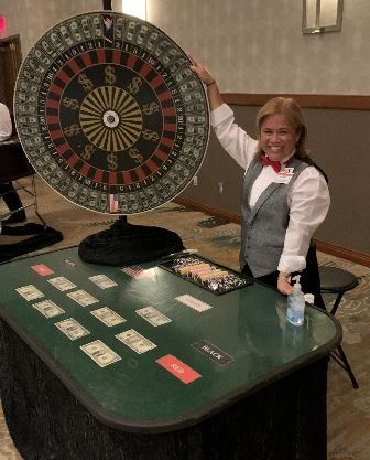 Woman at a casino roulette table, smiling and pointing at the wheel with chips and betting cards visible.
