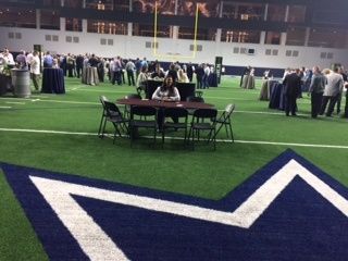 Indoor gym event with people standing around tables on green turf near a large blue-and-white star logo.