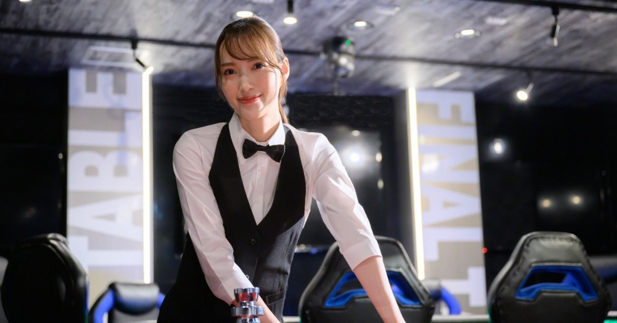 A woman casino dealer in a black vest and bow tie standing behind a gaming table with stacked chips.