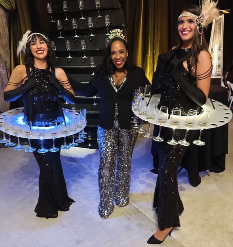 Three women in black outfits hold illuminated circular trays in a dim indoor venue.