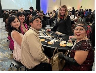 A group of people dressed in festive attire sits at a blackjack table in a ballroom, smiling at the camera.