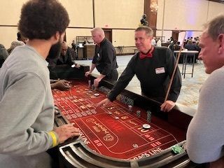 A craps table in a large room with a dealer pointing to the layout while players stand around and place bets.