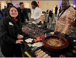 A smiling dealer at a roulette table in a crowded event hall, with onlookers standing around the game setup.