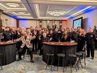 A group of people wearing black shirts and nametags cheers in a ballroom with casino tables, one person wears a patterned.